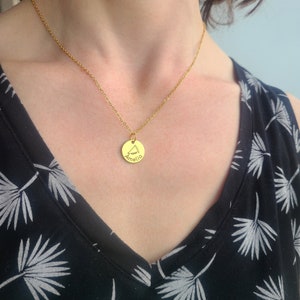 May include: A gold necklace with a round pendant that says "Amelia" on it. The necklace is being worn by a person with dark hair.