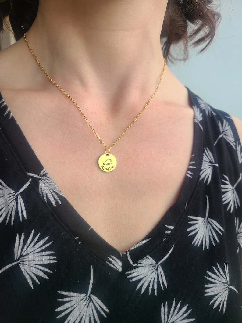 May include: A gold necklace with a round pendant that says "Amelia" on it. The necklace is being worn by a person with dark hair.