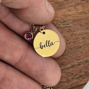 Op de afbeelding: Een goudkleurige ketting met een ronde hanger gegraveerd met de naam "bella" in cursief. Een kleine, ronde, paarse edelsteenbedel hangt naast de hanger. De ketting wordt in een hand gehouden.