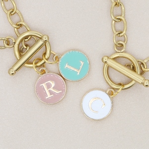 Puede incluir: Collares de cadena dorados con cierres de palanca. Cada collar presenta un dije redondo con una letra: rosa con "R", turquesa con "L" y blanco con "C". Los dijes están delineados en dorado.