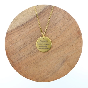 Puede incluir: Un collar dorado con un colgante circular. El colgante presenta una cita grabada de Ann Richardson: "We cannot change the outcome, but we can affect the journey." El collar se muestra sobre una superficie de madera.