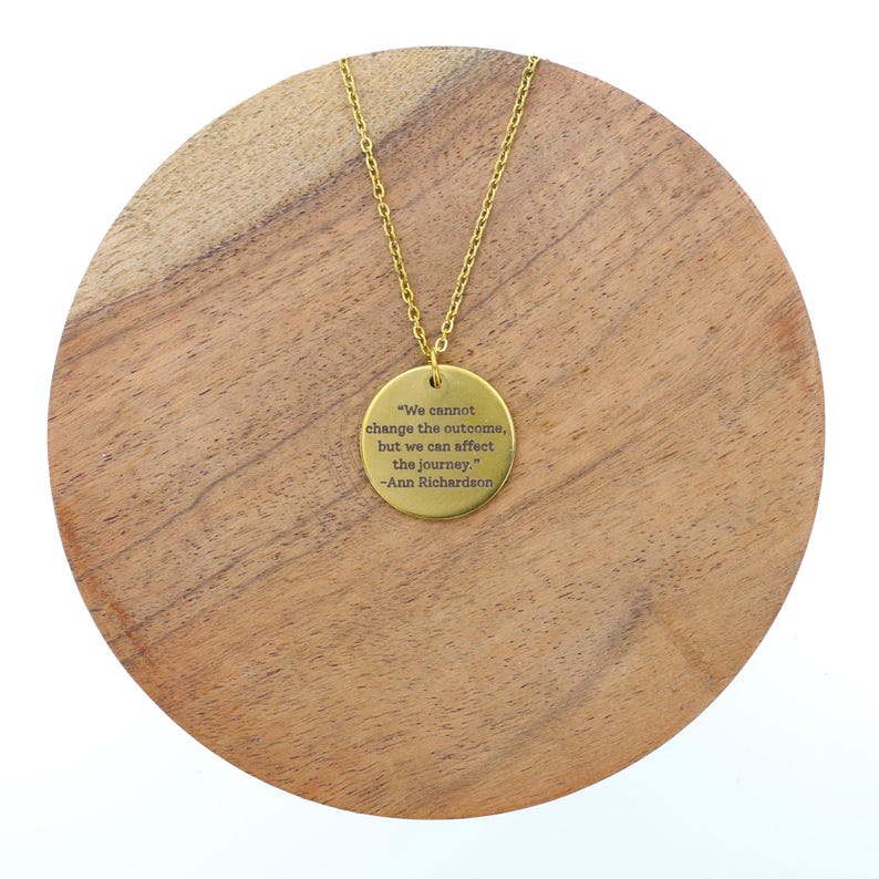 Puede incluir: Un collar dorado con un colgante circular. El colgante presenta una cita grabada de Ann Richardson: "We cannot change the outcome, but we can affect the journey." El collar se muestra sobre una superficie de madera.