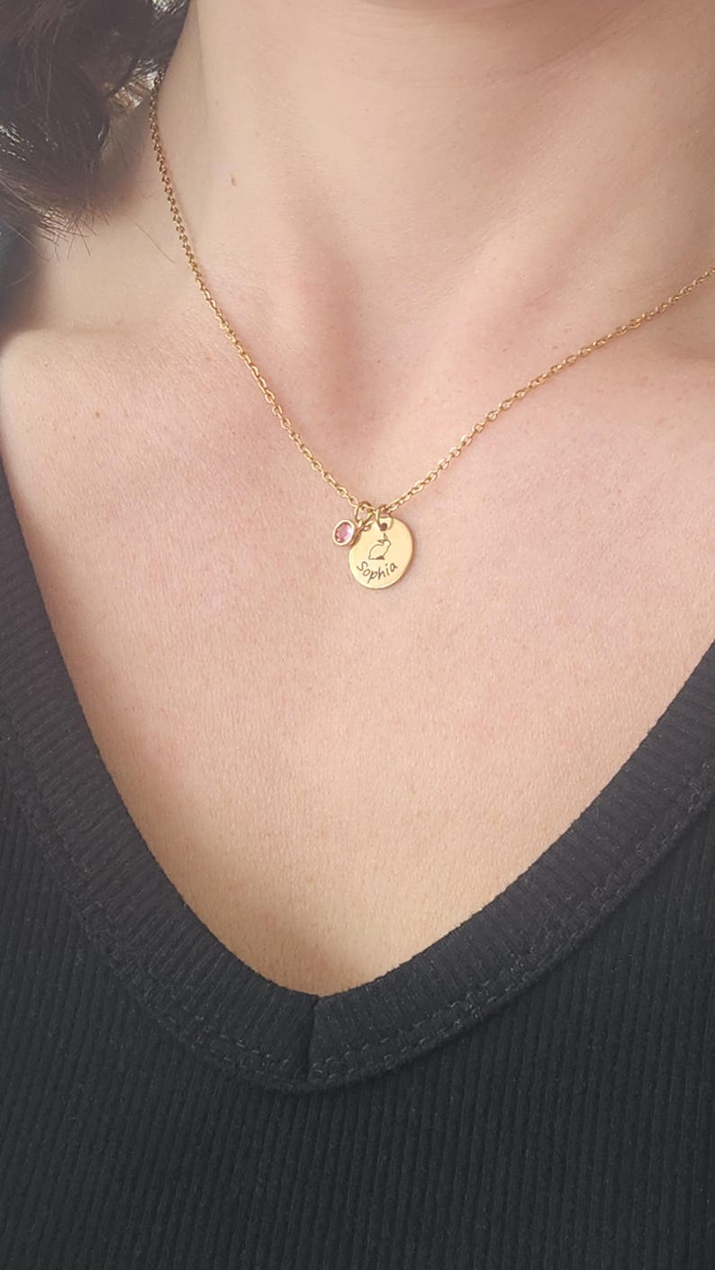 Model in black vneck shirt is wearing a gold necklace with pink crystal and small god disc engraved with bunny outline in the middle and name around the edge.
