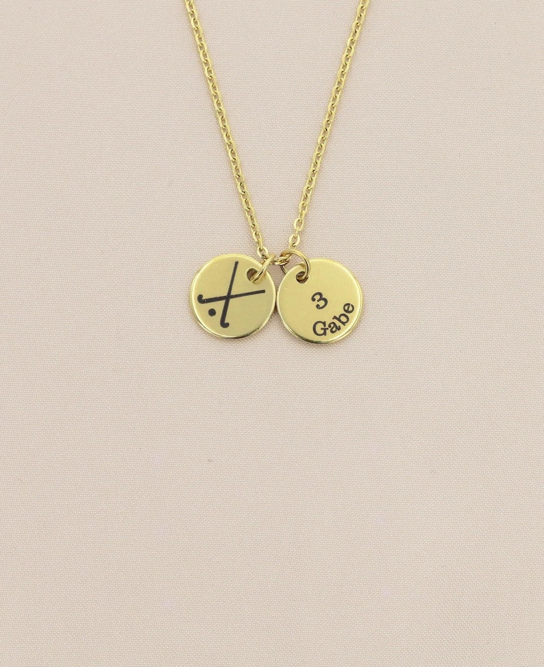gold necklace with two discs showing field hockey symbol and number name of player on cream background