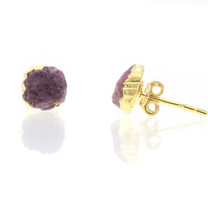 Pode incluir: Um par de brincos de pressão com pedras preciosas rosa escuro em bruto, em metal dourado. Os brincos têm uma aparência natural e orgânica, com as pedras preciosas exibindo uma superfície texturizada e formato irregular.