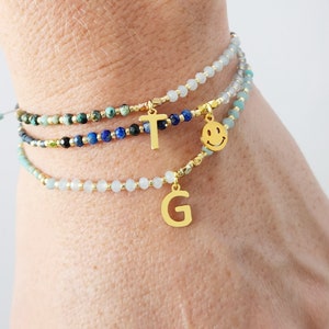 Può includere: Tre braccialetti a strati di perline con ciondoli dorati. Il braccialetto superiore ha perline turchesi e blu. Il braccialetto intermedio ha perline blu e nere. Il braccialetto inferiore ha perline blu chiaro. I ciondoli sono una lettera T dorata, una faccina sorridente dorata e una lettera G dorata.