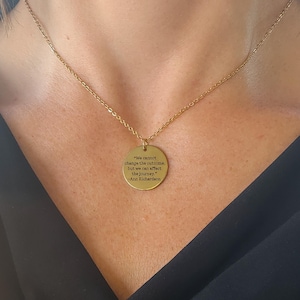 Puede incluir: Collar dorado con una cadena fina y un colgante redondo. El colgante lleva grabada la cita: "We cannot change the outcome, but we can affect the journey." -Ann Richardson. El collar se muestra sobre un fondo oscuro.