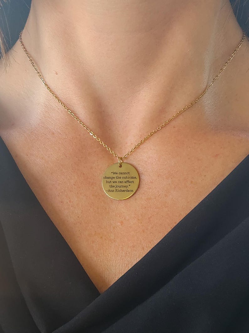 Puede incluir: Collar dorado con una cadena fina y un colgante redondo. El colgante lleva grabada la cita: "We cannot change the outcome, but we can affect the journey." -Ann Richardson. El collar se muestra sobre un fondo oscuro.