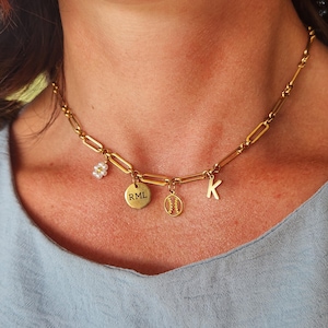 K&ouml;nnte beinhalten: Goldfarbene Kette mit mehreren Anh&auml;ngern. Zu den Anh&auml;ngern geh&ouml;ren eine kleine Blume, ein runder Anh&auml;nger mit der Gravur "RML", ein Baseball-Anh&auml;nger und der Buchstabe "K". Die Halskette wird von einer Person getragen.