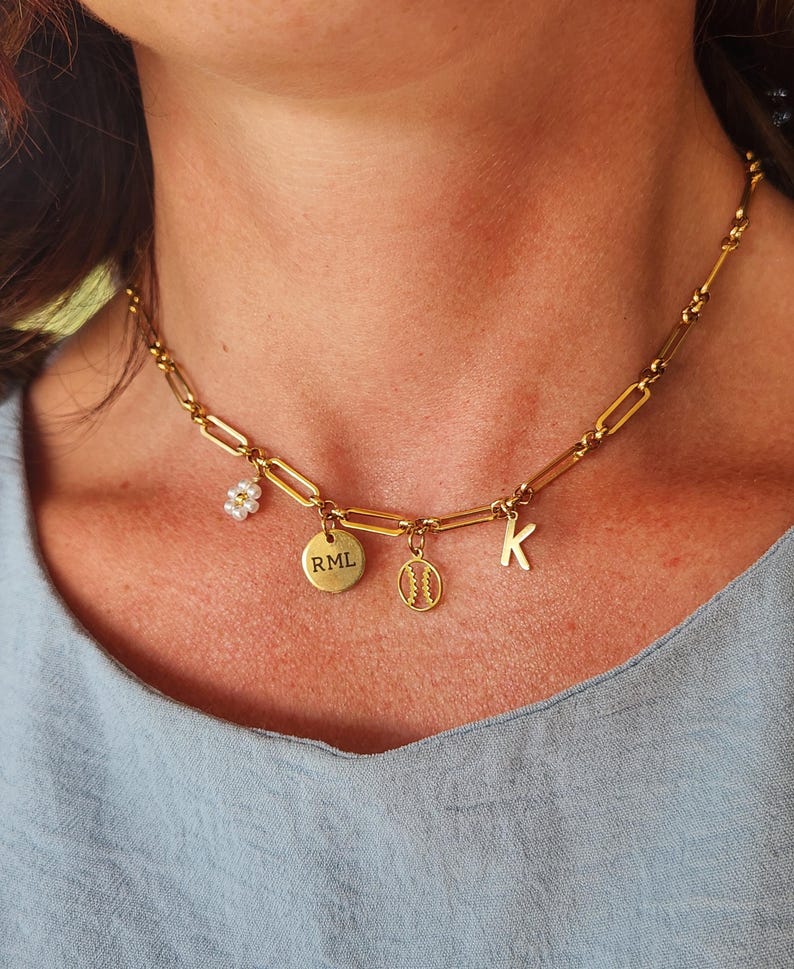 K&ouml;nnte beinhalten: Goldfarbene Kette mit mehreren Anh&auml;ngern. Zu den Anh&auml;ngern geh&ouml;ren eine kleine Blume, ein runder Anh&auml;nger mit der Gravur "RML", ein Baseball-Anh&auml;nger und der Buchstabe "K". Die Halskette wird von einer Person getragen.