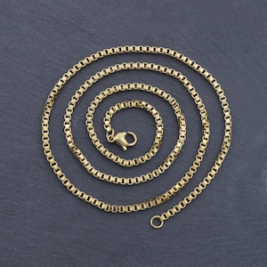 May include: A gold-colored box chain necklace, coiled in a spiral. The chain is made of small, rectangular links and features a lobster clasp closure. The necklace is displayed on a dark gray surface.