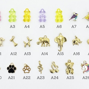K&ouml;nnte beinhalten: Eine Sammlung kleiner Charms in verschiedenen Formen und Farben. Die Charms umfassen Gummib&auml;rchen, V&ouml;gel, Tiere und Pfotenabdr&uuml;cke. Jeder Charm ist mit einer Buchstaben- und Zahlenkombination beschriftet.