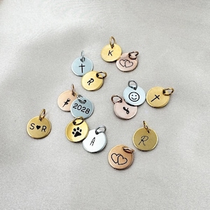May include: A collection of small, round metal charms in gold, silver, and rose gold tones. Each charm features a different design, including initials, symbols like a cross and paw print, and a smiley face. Some charms have a small loop for attaching to a chain.
