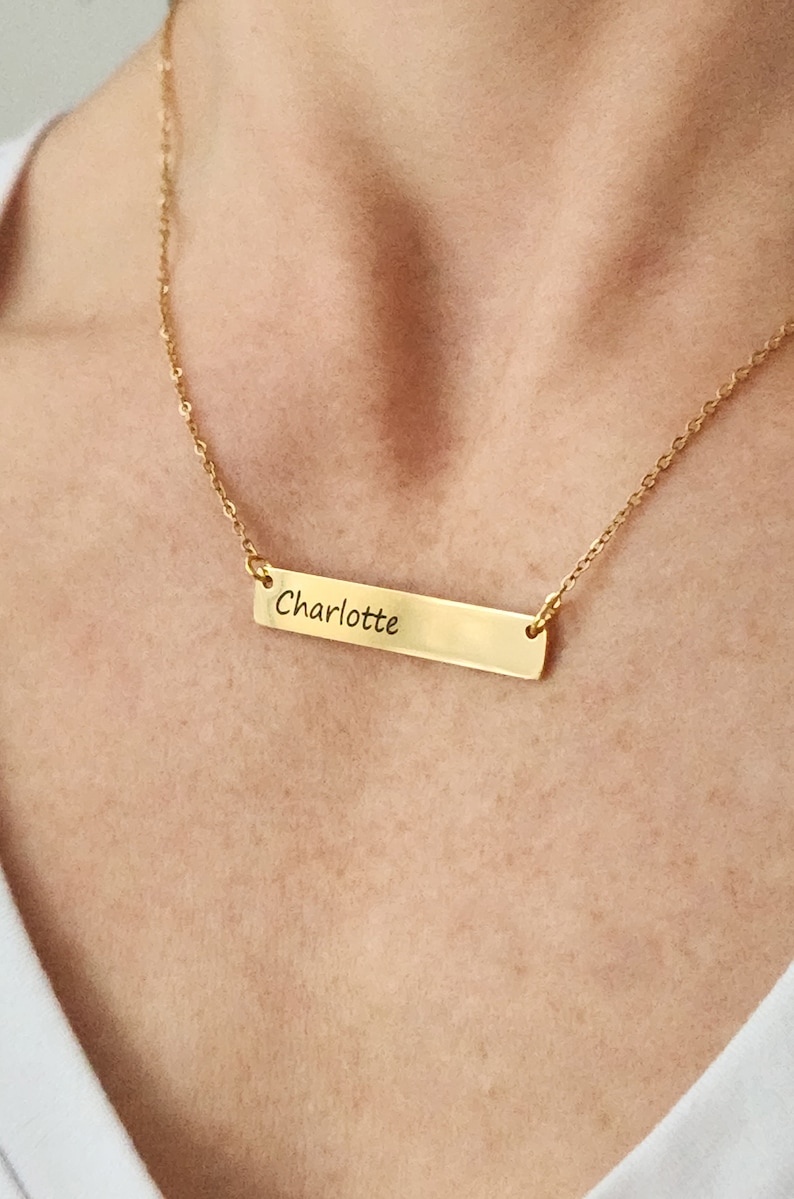 A female model  wearing a gold necklace with a rectangular bar pendant. The bar is engraved with the name "Charlotte."