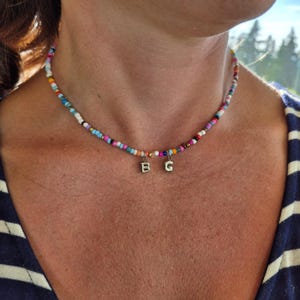 Puede incluir: Un collar de cuentas de colores con pequeñas cuentas en varios colores, incluyendo rosa, azul y blanco. El collar tiene dos pequeños colgantes plateados con las letras "B" y "G". El collar se lleva alrededor del cuello.