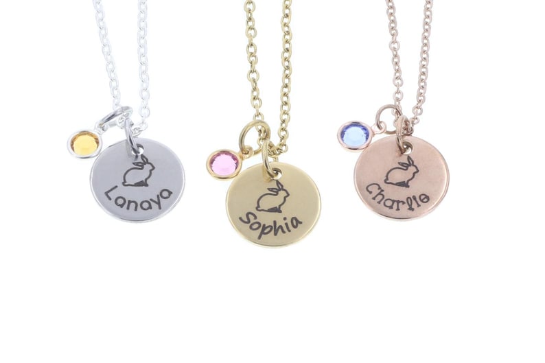 Three necklaces on white background. Each necklace has a small metal disc with a bunny outline engraved in the middle, a name around the edge, and colored crystal to the left. Each necklace is gold, rose gold, or silver.