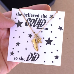 May include: A gold-toned necklace featuring a small soccer cleat charm and a letter K charm. The necklace is displayed on a white card printed with black stars and the text "she believed she COULD so she DID" in a mix of serif and script fonts.