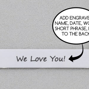 A silver rectangular pendant on a chain displays the engraved word "We Love You!" in black script. An arrow points to back, suggesting customizations: "Add engraved name, date, word, short phrase, etc. to the back!";