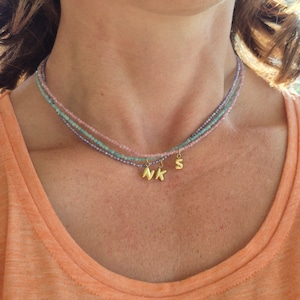 Pode incluir: Um colar em camadas com pequenas contas coloridas em tons de rosa, azul e roxo. Pingentes de iniciais douradas pendem do colar, exibindo as letras "N", "K" e "S".