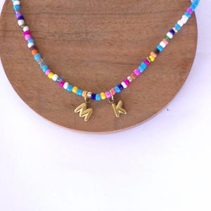 Puede incluir: Un collar de cuentas de colores con colgantes iniciales dorados. El collar presenta pequeñas cuentas redondas en tonos azules, rosas, amarillos y blancos. Los colgantes dorados tienen forma de las letras "M" y "K".