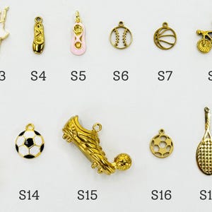K&ouml;nnte beinhalten: Eine Sammlung goldfarbener Charms in verschiedenen Formen, darunter Ballettt&auml;nzerinnen, Sportger&auml;te wie ein Football, ein Fu&szlig;ball und ein Tennisschl&auml;ger sowie andere Artikel. Jeder Charm ist mit einem Buchstaben und einer Nummer gekennzeichnet.