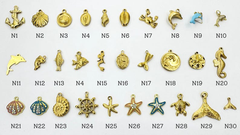 K&ouml;nnte beinhalten: Eine Sammlung goldfarbener nautischer Charms, darunter Anker, Muscheln, Delfine, Seesterne und Seepferdchen. Jeder Charm hat eine Schlaufe zum Befestigen an Schmuck oder anderen Bastelarbeiten. Die Charms sind in Reihen und Spalten angeordnet, wobei jeder Charm mit einer Nummer versehen ist.
