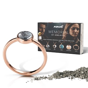 May include: A rose gold ring with a round, gray stone. A box labeled "Memorial DIY Jewelry Kit" is in the background, along with a pile of gray material. The kit includes options for wearable memorials.