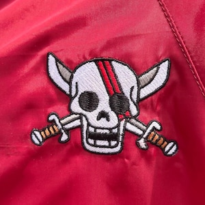 May include: Red embroidered patch with a white skull and crossbones. The skull has a black eye patch and a red stripe across the forehead. The crossbones are made of two silver swords.