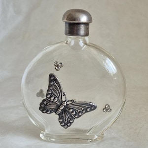 Puede incluir: Una botella de vidrio transparente con un diseño de mariposa plateada y una tapa plateada. La botella es redonda y tiene una pequeña abertura en la parte superior.
