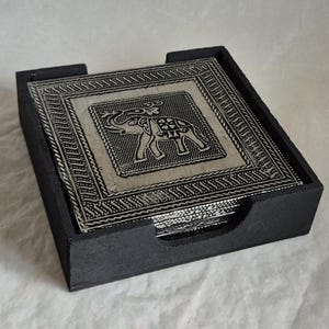 May include: A set of square coasters with an elephant design in a black holder. The coasters have a silver-coloured centre with an embossed elephant and a decorative border. The holder is black and has a slot for easy access to the coasters.