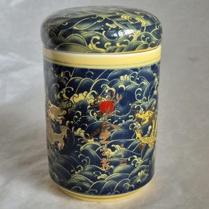 May include: A cylindrical ceramic container with a lid. The container is navy blue with a yellow band and features a design of golden dragons, red suns, and stylized waves. The lid mirrors the wave pattern.