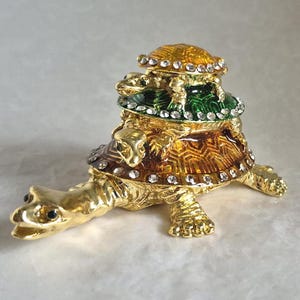 Può includere: Statuetta a forma di tartaruga dorata con una pila di tre gusci di tartaruga sulla schiena. I gusci sono decorati con smalto verde, marrone e giallo e impreziositi da piccoli cristalli trasparenti. La tartaruga ha occhi neri ed è appoggiata su una superficie bianca.