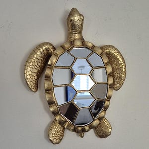 Könnte beinhalten: Ein goldfarbener, schildkrötenförmiger Wandspiegel mit einem Mosaik aus kleinen, verspiegelten Quadraten.