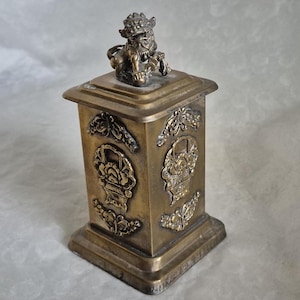 Puede incluir: Una caja de metal dorado con intrincados diseños florales y un león en la parte superior. La caja es cuadrada y tiene una base elevada.