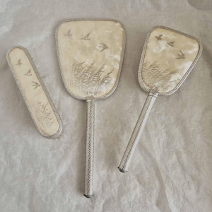 Vintage 3 piece vanity set, attractive decoration depicts birds in flight amidst tall grasses; mirror, hairbrush and clothes brush.