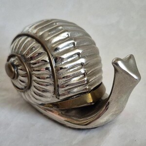 Puede incluir: Un pisapapeles plateado con forma de caracol. La concha del caracol tiene una textura acanalada y el cuerpo es liso. El diseño incluye una banda de color latón alrededor de la concha. La cabeza del caracol está extendida.