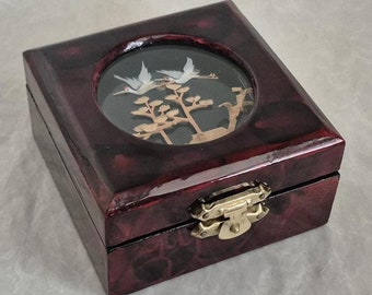 May include: A small, square, wooden box with a hinged lid and a brass latch. The box is painted a deep red with a marbled effect. A clear glass window on the top of the box reveals a delicate paper craft of two white birds flying over a tree.