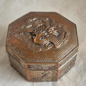 May include: Octagonal shaped, copper coloured trinket box with an embossed design of a phoenix on the lid. The box has a textured surface.