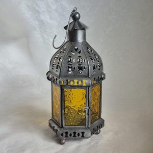 May include: A hexagonal metal lantern with a dark grey finish and amber-coloured glass panels. The lantern features intricate cut-out patterns on the top and base, a hinged door with a small latch, and a curved metal handle at the very top.