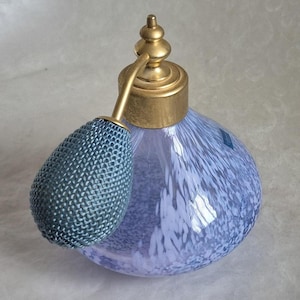 Puede incluir: Un frasco de perfume vintage con un cuerpo de vidrio moteado azul claro y blanco. Cuenta con una tapa y un atomizador de metal dorado, y una pera de atomizador azul a juego. El frasco tiene un diseño clásico y elegante.