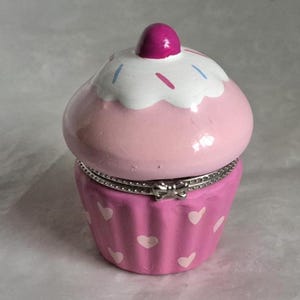 May include: A pink cupcake-shaped trinket box with a white frosted top and a magenta cherry. The cupcake base is a darker pink with white heart accents. The box is hinged with a silver-coloured metal band.
