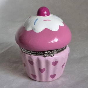 May include: A pink cupcake-shaped trinket box with a hinged lid. The top is decorated with white frosting and a cherry. The base is light pink with heart-shaped accents. A silver-colored metal band and bow detail the hinge.