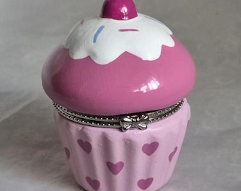 May include: A pink cupcake-shaped trinket box with a hinged lid. The top is decorated with white frosting and a cherry. The base is light pink with heart-shaped accents. A silver-colored metal band and bow detail the hinge.