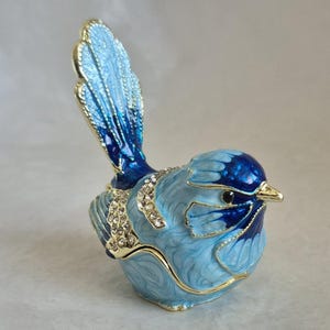 May include: A bird-shaped trinket box featuring light blue and dark blue enamel with gold-tone metal trim. The figurine is decorated with clear rhinestones along the wing and chest area, and it has a small, pointed gold-tone beak and black eyes.