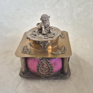 Puede incluir: Una caja decorativa de plata y oro con un interior rosa. La tapa presenta una escultura de león de plata detallada.