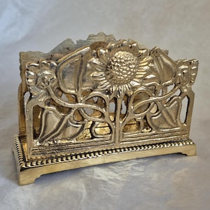 May include: A gold-colored metal napkin holder with a sunflower design. The holder features an intricate floral pattern with a detailed sunflower in the center and decorative elements on the sides. The base has a beaded trim.