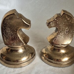 May include: Two gold-colored chess knight figurines. Each knight is sculpted with a detailed horse head design, mounted on a round base. The figurines have a metallic finish and are displayed on a white surface.