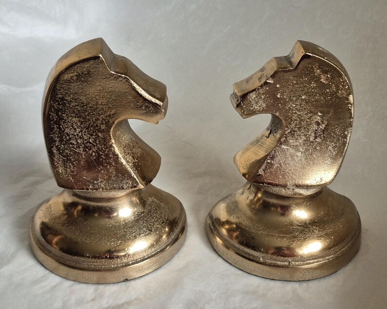 May include: Two gold-colored chess knight figurines. Each knight is sculpted with a detailed horse head design, mounted on a round base. The figurines have a metallic finish and are displayed on a white surface.