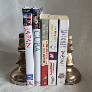May include: A set of books held between two gold-colored chess piece bookends. The books include titles like "Japan," "Cuba," and "The Curious Incident of the Dog in the Night-Time."