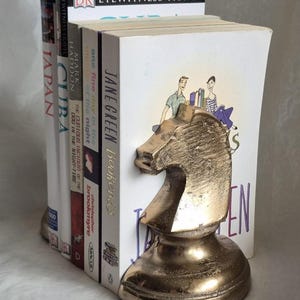 May include: A gold-colored metal knight chess piece bookend holding a stack of books. The books have titles like "Japan", "Cuba", and "Jane Green Bookends". The bookend is a decorative item for home or office.